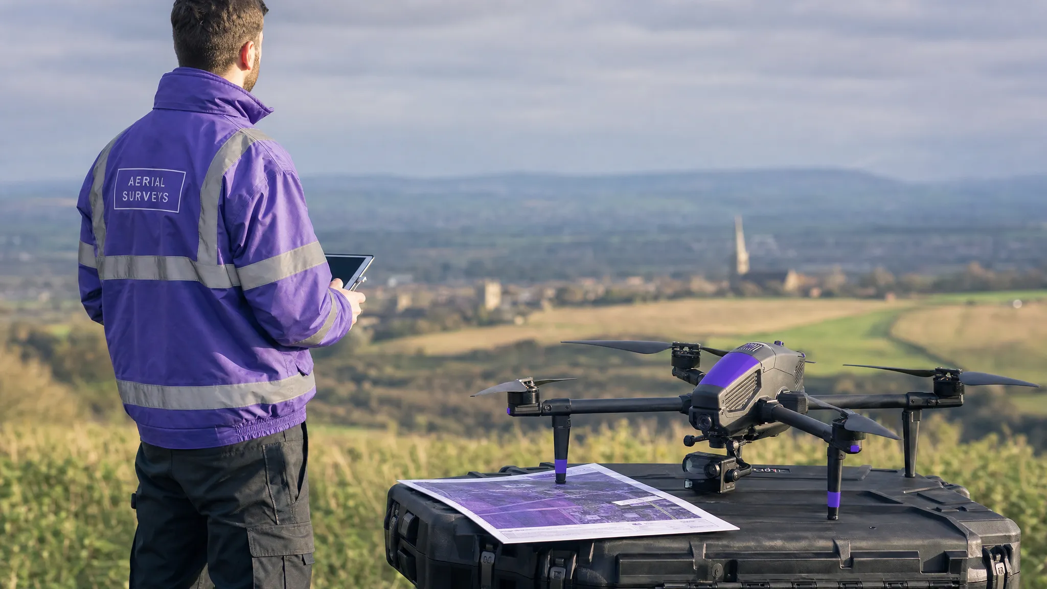 A commercial drone resting on a hard case beside a printed site plan in a UK field, with a pilot checking surrounding landmarks before a planned survey flight.