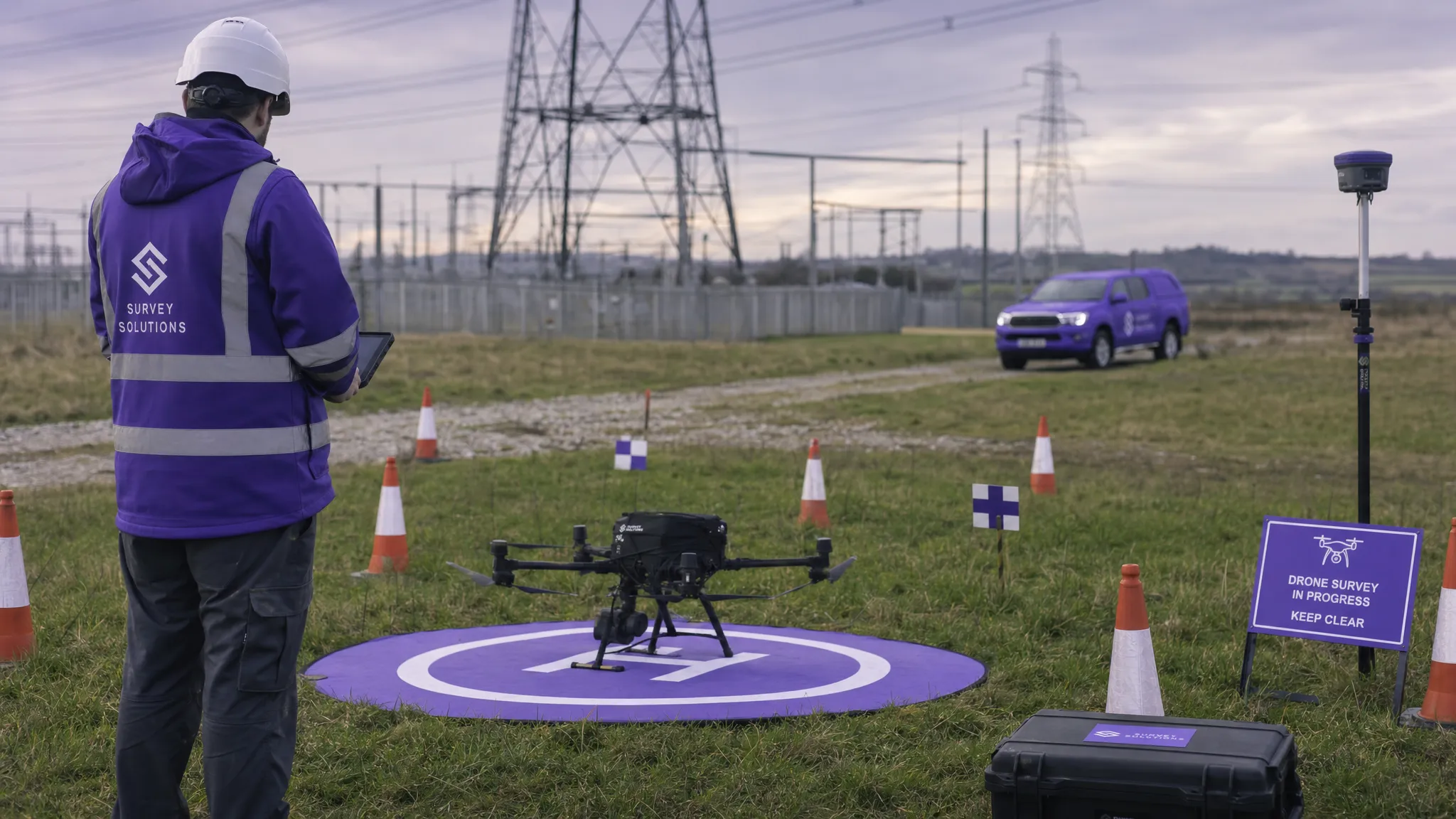 A drone survey team setting up a safe take-off area in an open field near an infrastructure site, with cones marking the operating area, a UAV on a landing mat, survey markers on the ground and pylons and access tracks visible in the distance.